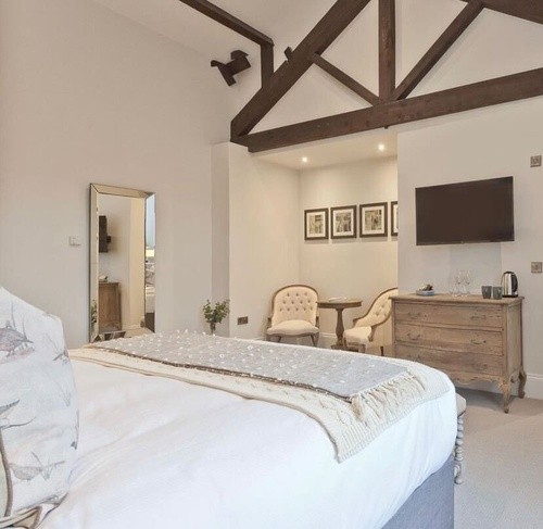 made bed with white comforter in a tall room with wood beams in the ceiling and white chairs in the corner